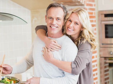 A man and woman smiling and embracing in a kitchen while preparing a healthy meal together